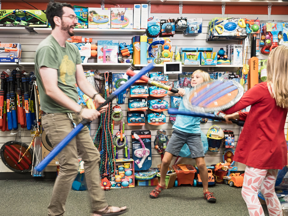 An adult and two older children pretend sword fighting in a toy store